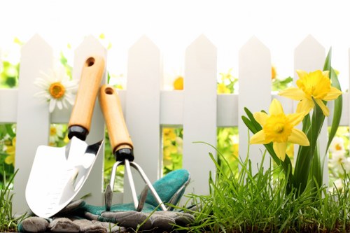 The image depicts a garden scene in front of a white picket fence with a blurred background of lush greenery and flowers. In the foreground, there are two gardening trowels with wooden handles and metal blades, leaning against each other. The trowels are placed on dark soil or mulch, surrounded by green grass and small yellow daffodil flowers with long green leaves. The grass appears dense and well-maintained, and the overall scene suggests a neatly cared-for outdoor space suitable for gardening and landscaping services in Blackwall and the nearby area, emphasizing tools and plant elements typical of garden maintenance work. The weather appears bright with natural sunlight illuminating the scene, creating a fresh and inviting atmosphere consistent with professional garden care environments.