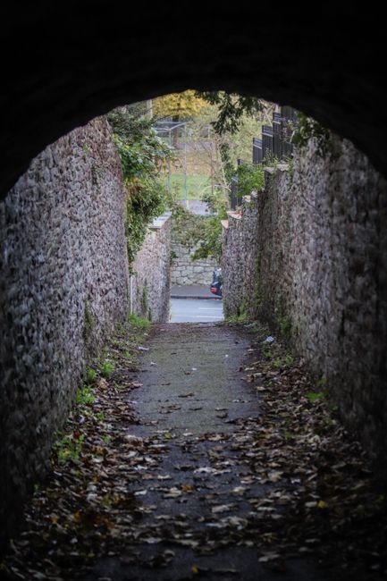 A narrow, slightly sloping garden pathway viewed from beneath an arched brick bridge, with both sides bordered by weathered stone walls covered in patches of moss and climbing greenery. The ground is